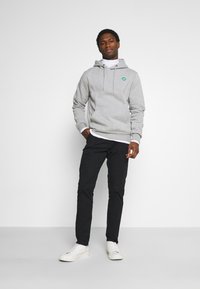 Sudadera gris con cordones y logo verde, superpuesta sobre un cuello de tortuga blanco, combinada con pantalones negros y zapatillas blancas.