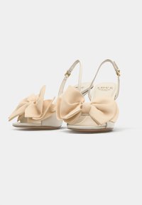 Μπεζ ανοιχτά σανδάλια με γυαλιστερή φινίρισμα, που διαθέτουν μεγάλες υφασμάτινες κορδέλες μπροστά. Ρυθμιζόμενοι ιμάντες αστραγάλου με χρυσές λεπτομέρειες.