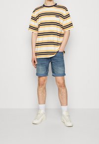 Camiseta de rayas en amarillo, azul marino y blanco con un corte relajado. Combinada con pantalones cortos de denim enrollados y zapatillas blancas. Detalle de marca sencillo.
