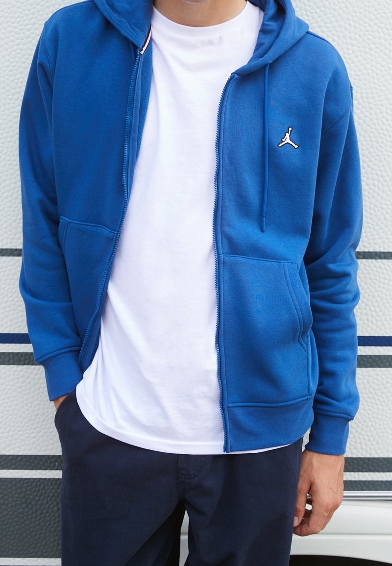 Personne portant un sweat à capuche bleu zippé par-dessus un t-shirt blanc et un pantalon foncé, debout devant un fond texturé blanc et gris.