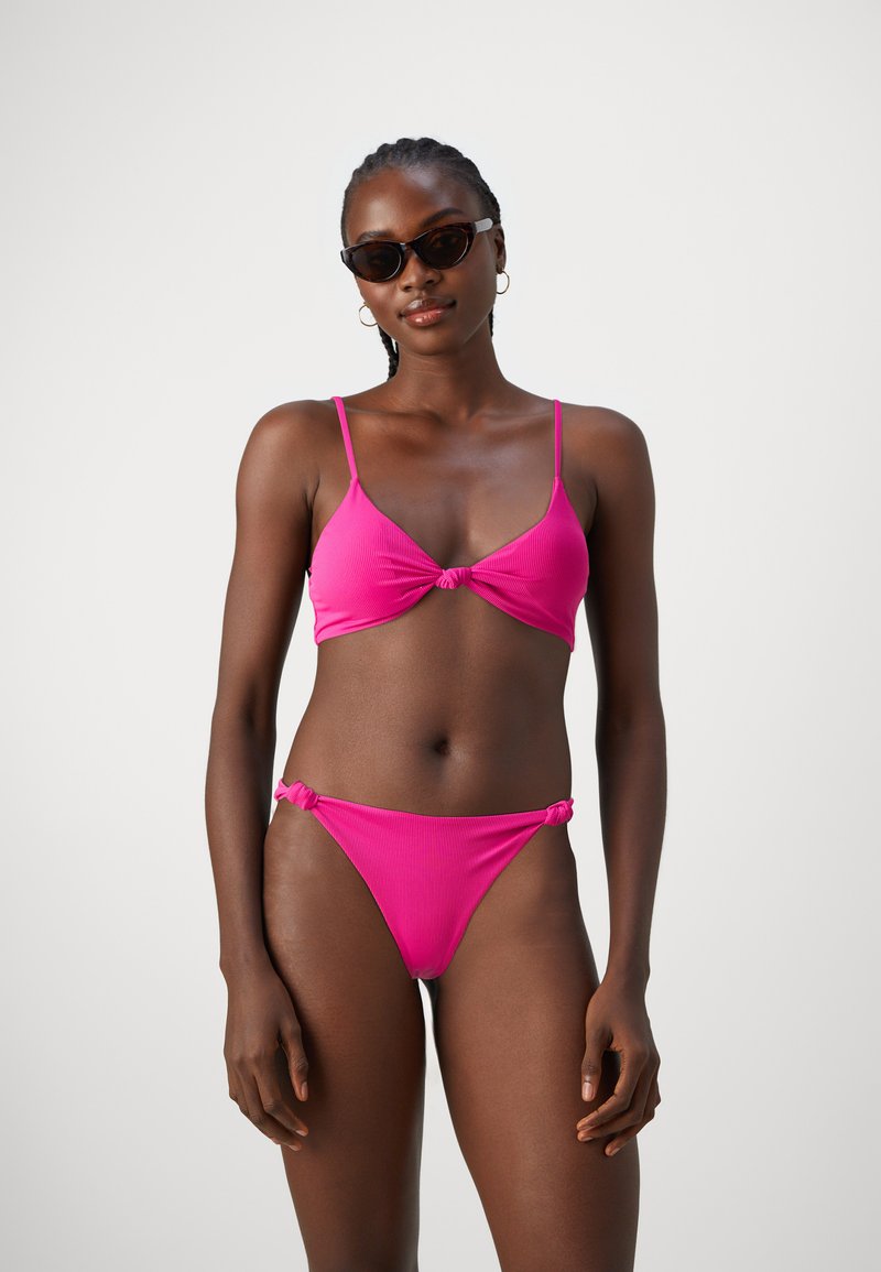 Rózsaszín bikini szett, amely csomózott kialakítással, vékony pántokkal és bordázott textúrával rendelkezik, valamint egy hozzáillő alsó résszel. Sötét napszemüveget is tartalmaz kiegészítőként.