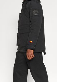 Veste noire isolée avec capuche, dotée de poches zippées et à rabat. Logo brodé sur la manche et fermetures à boutons à l'avant.