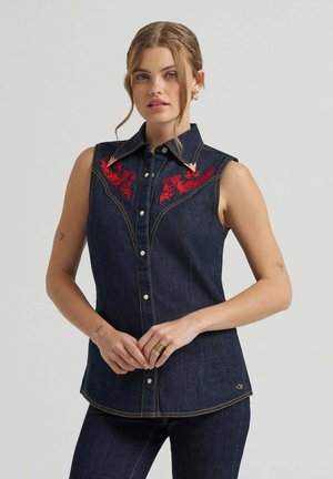 Frau trägt ärmelloses dunkelblaues Jeanshemd mit roter Blumenstickerei auf der Brust, Knopfleiste vorne und Kragen, steht mit gefalteten Händen.