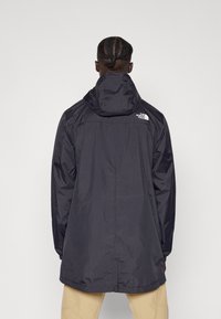 Svart huva regnjacka med mattsl finish, lång design och vitt logotyp på ryggen. Har en avslappnad passform och avslappnad sömnad.