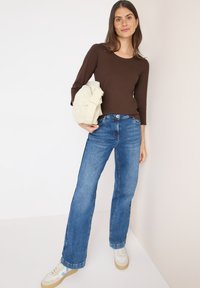 Maglietta marrone a manica lunga abbinata a jeans dritti blu chiaro. Il modello tiene una borsa beige e indossa sneakers bianche e blu.