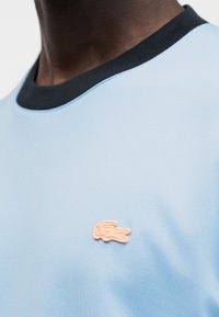 T-shirt bleu clair avec un col côtelé noir et un petit logo rose brodé sur la poitrine. Texture de tissu lisse, design décontracté.