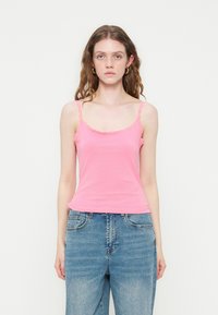 Jeune femme aux cheveux bruns ondulés portant des boucles d'oreilles créoles dorées, un débardeur rose avec bordure en dentelle, et un jean en denim bleu, debout devant un fond blanc.