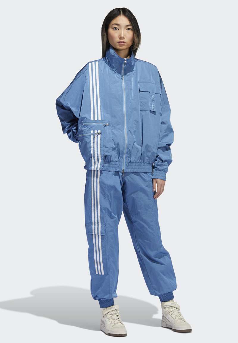 Lichtblauwe trainingsoutfit met een ritsjack en elastische boorden. Witte strepen lopen langs de zijkanten; inclusief zakken en een hoge kraag.