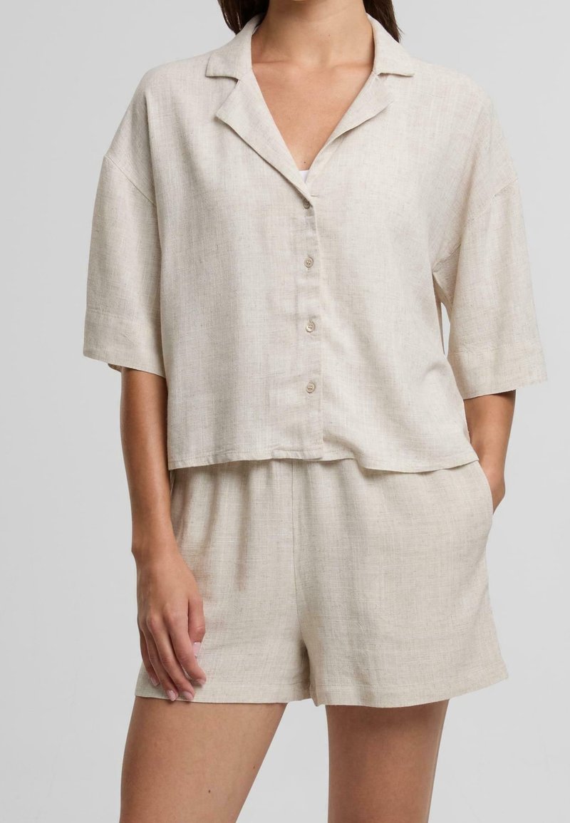 Femme portant une chemise beige en lin à manches courtes boutonnée et un short ample assorti, une main dans la poche, sur fond gris.