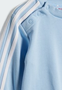 Felpa azzurra chiara in cotone, con strisce bianche sulle spalle e bottoni a pressione vicino al collo. Texture morbida.