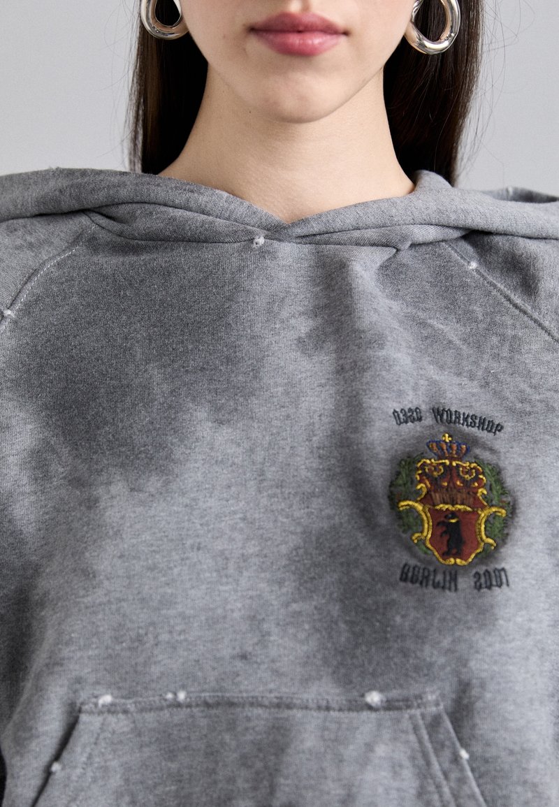 Jeune femme portant des boucles d'oreilles créoles argentées et un sweat à capuche gris avec un petit blason brodé sur la poitrine.