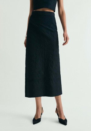 Mujer con falda midi negra texturizada, top corto sin mangas a juego y tacones slingback negros de punta puntiaguda, de pie sobre un fondo liso.