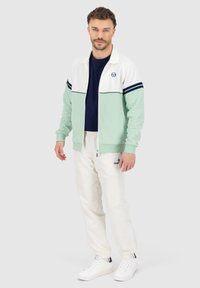 Mintgrønn og hvit zip-up jakke med marineblå striper, kombinert med hvite bukser og hvite sneakers, med en logo på jakken og buksene.