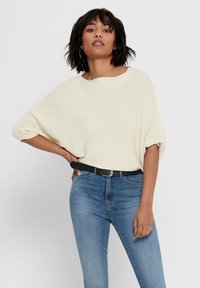 Cream-colored, ribbed knit oversized sweater with wide sleeves, paired with blue denim jeans and a black belt. Simple, relaxed design.