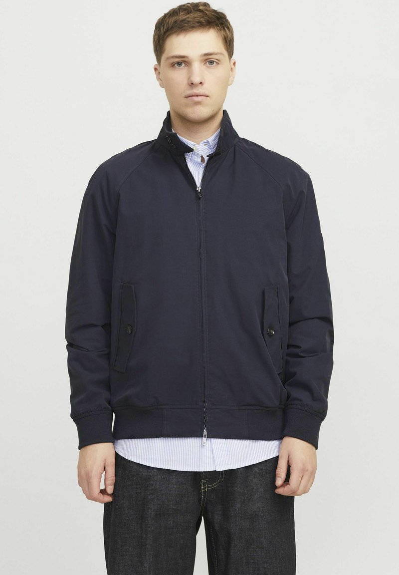 Navy pullover jacket with a front zipper, stand collar, side pockets, and ribbed cuffs, paired with a blue and white striped shirt.