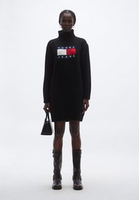 Musta neulemekko, jossa on korkea kaulus ja etuosassa punaisen, valkoisen ja sinisen värinen logo. Malli pitää kädessään pientä mustaa käsilaukkua ja pukeutunut ruskeisiin saappaisiin.