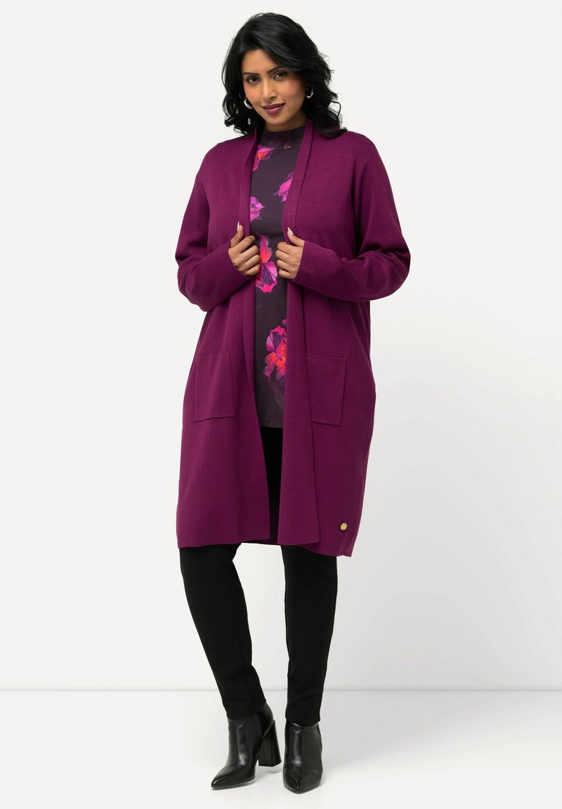 Langer lila Cardigan aus weichem Stoff, mit offenem Frontdesign, zwei vorderen Taschen und einem goldenen Akzent am Saum.