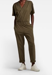 Olivgrön jumpsuit med omslagdesign, korta ärmar, elastisk midja och tapered byxor, kombinerad med vita sneakers. Slät tygtextur.