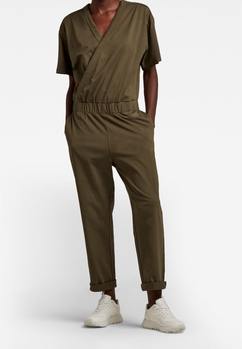 Olivgrön jumpsuit med omslagdesign, korta ärmar, elastisk midja och tapered byxor, kombinerad med vita sneakers. Slät tygtextur.