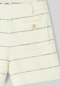 Shorts en tissu à texture crème avec de fines rayures noires, une poche à rabat à l'avant avec un bouton doré, et une petite étiquette blanche à la taille.