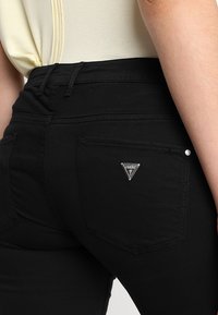Schwarze Denim-Jeans mit figurnahem Schnitt, versehen mit einem silbernen Dreieckslogo-Akzent auf der Gesäßtasche. Glatte Textur und klassischer Fünf-Taschen-Stil.
