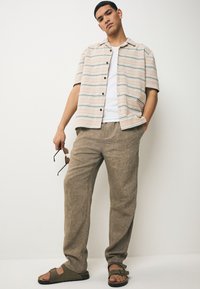 Camicia a maniche corte a righe beige con linee blu; abbinata a pantaloni larghi color sabbia e sandali marroni. Accessori includono occhiali da sole.