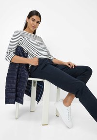 Gestreiftes Langarmshirt, marinefarbene Hose und weiße Sneaker. Eine marinefarbene gesteppte Jacke hängt über einem weißen Stuhl. Minimalistischer Hintergrund.