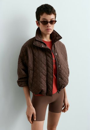 Personne aux cheveux courts porte une veste matelassée marron, une chemise rouge, un short marron ajusté et de petites lunettes de soleil foncées, debout devant un fond uni.