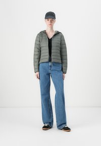 Polstret olivengrønn jakke med hette, sammen med en svart topp og blå vide jeans. Svart caps og joggesko fullfører antrekket.