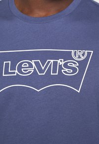 Námořnická modrá bavlněná tričko s bílou grafikou loga Levi's, která má zakřivený design na přední straně. Krátké rukávy a kulatý výstřih.