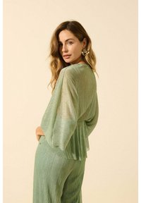 Tenue plissée vert clair avec de larges manches et une taille peplum. Texture transparente avec un subtil éclat. Accessoirisée de grosses boucles d'oreilles décoratives.