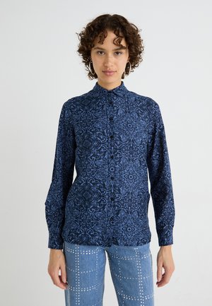 Camicia blu navy con intricati motivi blu chiaro, dotata di colletto e maniche lunghe. Indossata con jeans blu chiaro con borchie.