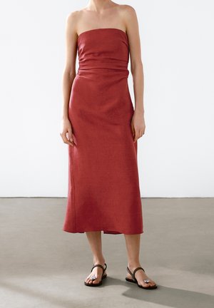 Vrouw draagt een strapless midi-jurk in roestrood en zwarte sandalen, staand op een grijze vloer tegen een witte muur.