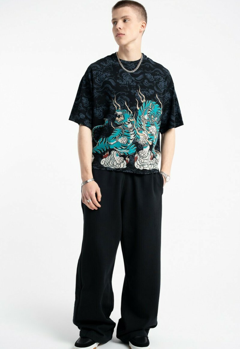 Młody mężczyzna ubrany w czarną, przewlekłą koszulkę z niebieskimi wzorami tygrysa, czarne szerokie spodnie, srebrny łańcuszek na szyi, bransoletkę, pierścionki i sneakersy.