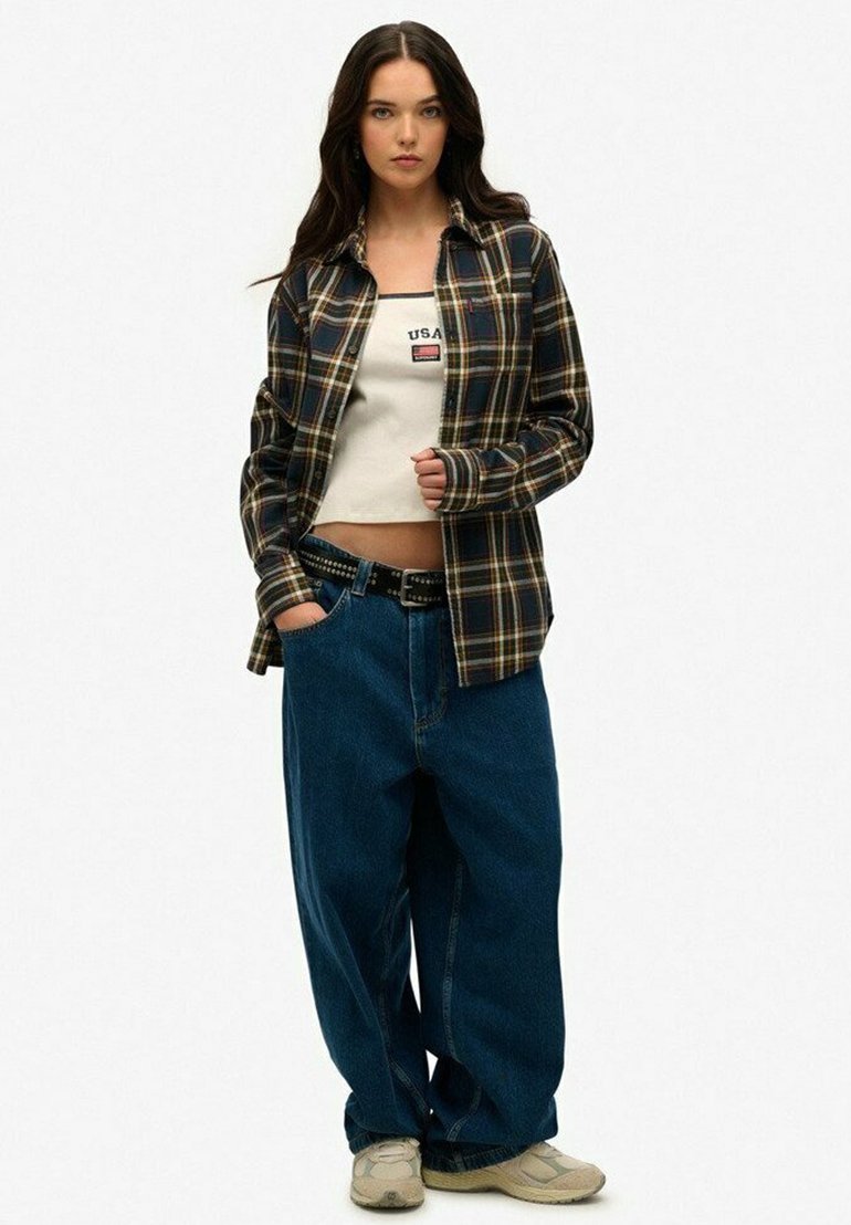 Chemise à carreaux bleu marine et jaune, superposée à un crop top blanc, associée à un jean bleu ample et des baskets beiges.