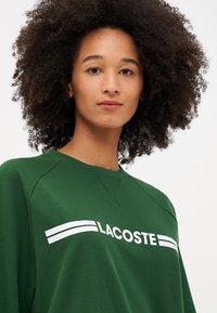 Groene trui met wit 'LACOSTE' logo, voorzien van een ronde hals en raglanmouwen, gemaakt van een gladde katoenmix.