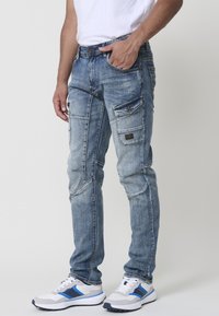 Jeans de denim azul claro con múltiples bolsillos, áreas desgastadas y detalles cosidos. Llevados con zapatos deportivos blancos y azules.