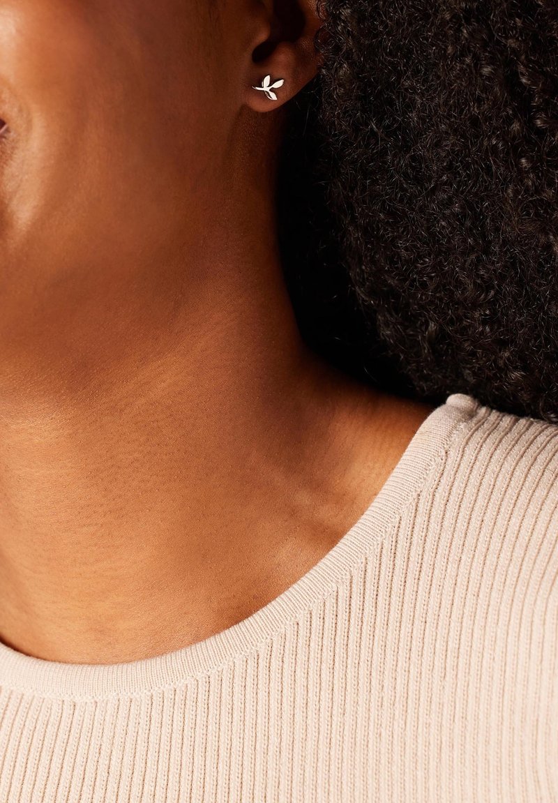 Zilveren oorstud in de vorm van een blad met een gepolijste afwerking, geplaatst tegen een huidskleur, aangevuld met de licht geribbelde halslijn van een trui.