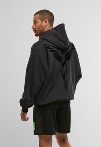 Mann in einem schwarzen Kapuzenpullover mit großem geflügeltem Figuren-Design auf dem Rücken, trägt schwarze Shorts mit einem grünen und gelben Abzeichen an der Seite.