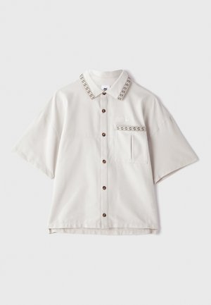 Camisa de manga corta con cuello en tejido beige claro. Presenta botones marrones, un bolsillo con tapa y un ribete de cuello con patrón.