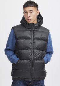 Schwarze Steppweste mit Kapuze, ausgestattet mit einem Reißverschluss, gestepptem Design und kleinem Logo auf der Vorderseite. Getragen über einem blauen Langarmshirt.