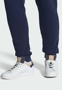 Valged tennisjalatsid, millel on augustamisdetailid, beežid aktsendid kontsapooles, kombineeritud tumesiniste spordipükstega, millel on elastsed mansetid.