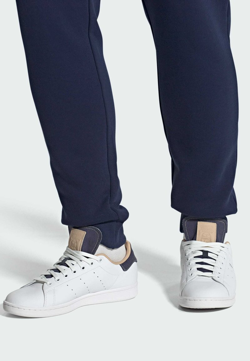 Valged tennisjalatsid, millel on augustamisdetailid, beežid aktsendid kontsapooles, kombineeritud tumesiniste spordipükstega, millel on elastsed mansetid.