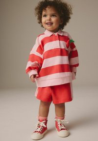 Polo surdimensionné à rayures rouges et roses avec un accent en forme de fraise, associé à un short rouge et des baskets montantes rouges. Matériel en coton doux.