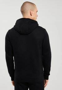 Bărbat văzut din spate, purtând un hanorac negru simplu cu gluga lăsată în jos și pantaloni negri asortați, pe un fundal deschis simplu.