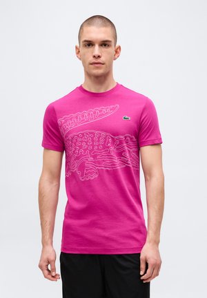 Homme portant un t-shirt rose vif avec un grand dessin en contour d’alligator et un petit logo de crocodile vert sur la poitrine.