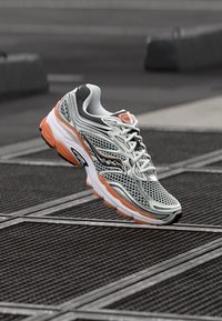 Leichter Laufschuh mit einem grauen Mesh-Obermaterial, orangen Akzenten und einer gepolsterten weißen Sohle. Besitzt ein atmungsaktives Design und eine strukturierte Oberfläche.