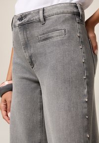 Pantalones de mezclilla gris con un corte ajustado, bolsillos laterales y una fila decorativa de remaches metálicos a lo largo de la costura exterior. Textura sutil visible.