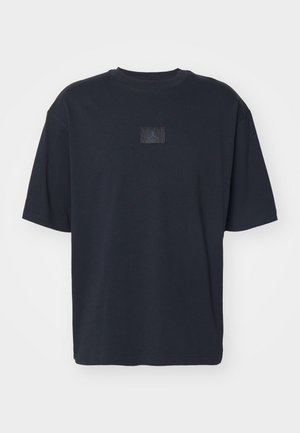 Camiseta de algodón negra con cuello redondo, mangas cortas y un parche de logo texturizado en el frontal. Corte simple y cuadrado, sin estampados.