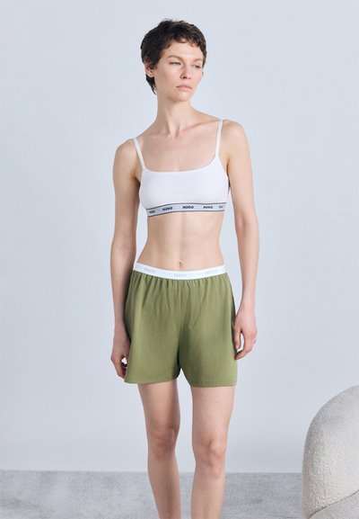 Femme aux cheveux courts portant un soutien-gorge de sport blanc et un short vert olive, debout devant un fond gris clair uni.